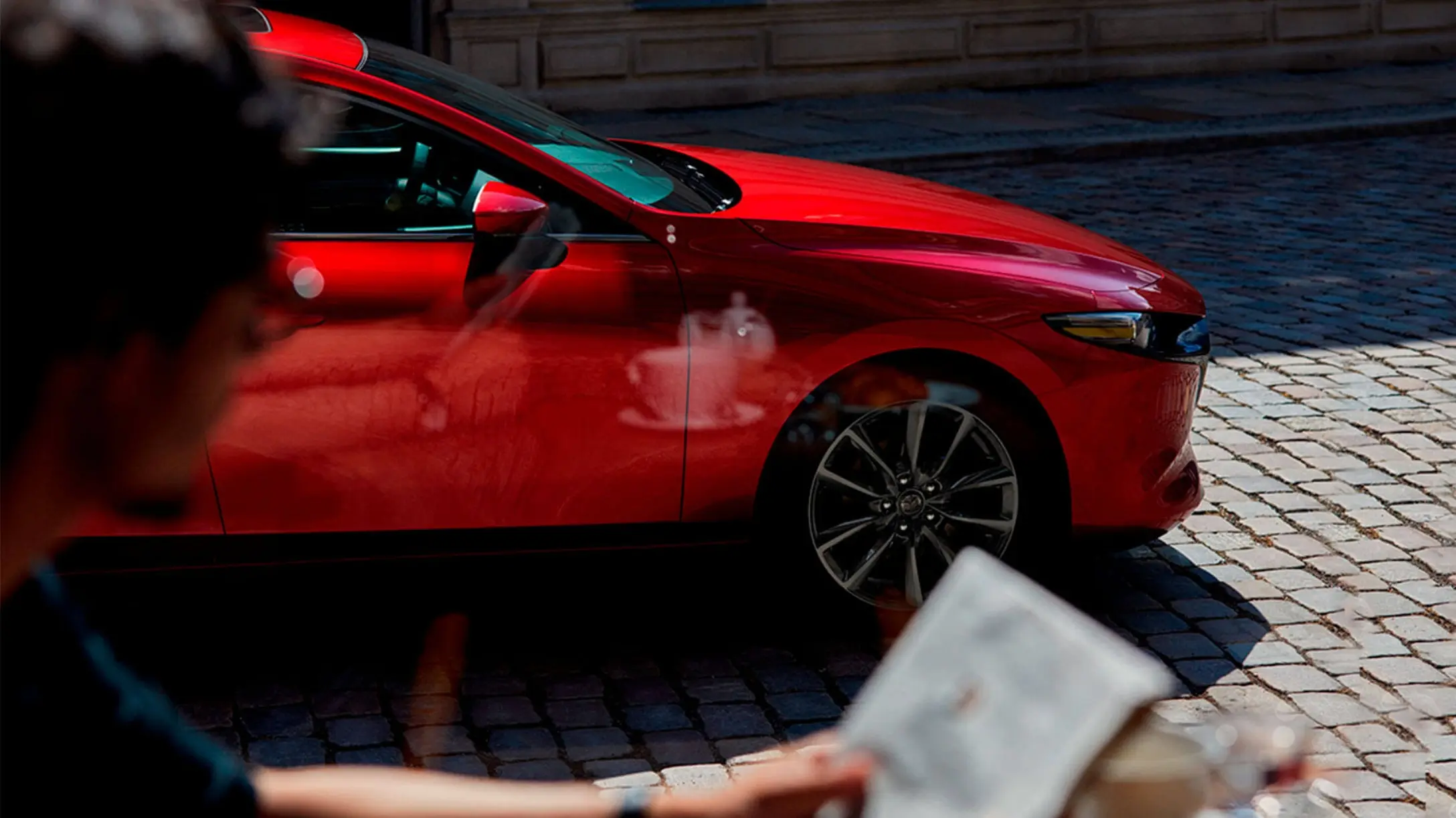 Mazda 3 Sport rojo estacionado en una calle de adoquines, visto desde un café.